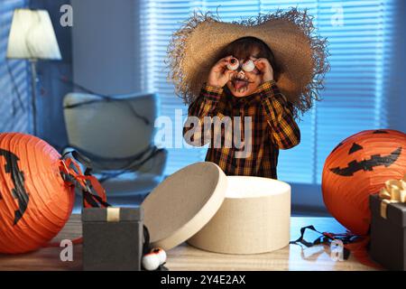 Süßer Junge, der wie Vogelscheuche gekleidet ist, mit dekorativen Augäpfeln, festlicher Dekoration und Geschenkboxen drinnen in der Nacht. Halloween-Feier Stockfoto