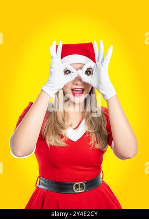 Die junge Latina-Frau, die als weihnachtsmann verkleidet ist, schaut in die Kamera und legt ihre Hände in ein OK-Schild, das ihre Augen mit gelbem Hintergrund gläsert Stockfoto