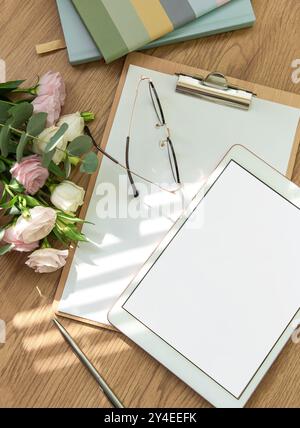 Ein heller Arbeitsbereich mit leerem Tablet, offenem Notizbuch, Schreibutensilien und frischen Blumen auf einem Holztisch lädt zu Kreativität und Kreativität ein Stockfoto