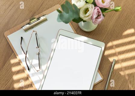 Ein heller Arbeitsbereich mit leerem Tablet, offenem Notizbuch, Schreibutensilien und frischen Blumen auf einem Holztisch lädt zu Kreativität und Kreativität ein Stockfoto