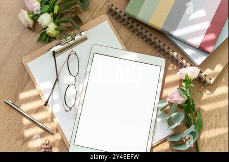 Ein heller Arbeitsbereich mit leerem Tablet, offenem Notizbuch, Schreibutensilien und frischen Blumen auf einem Holztisch lädt zu Kreativität und Kreativität ein Stockfoto