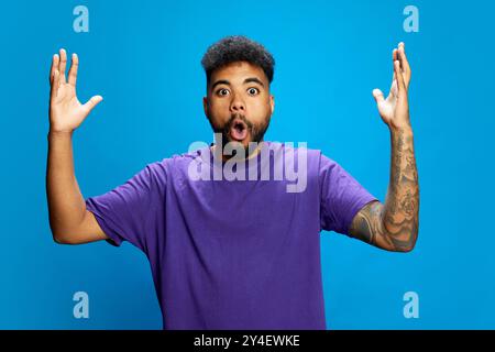 Ein afroamerikanischer Mann, der ein lila Hemd trägt, sieht überrascht aus, als er den Mund offen hält und seine Hände in verspielter Geste vor blauem Hintergrund hebt. Stockfoto