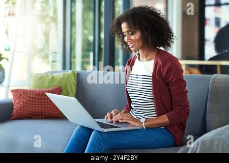 Schwarze Frau, zu Hause und Lächeln auf Sofa mit Laptop für Fernarbeit als freischaffender Schriftsteller oder Blogger. Weibliche Person, Angestellte und glücklich als Journalistin Stockfoto