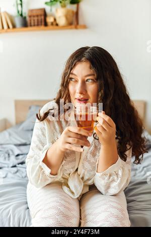 Eine schöne junge Frau sitzt auf ihrem Bett, schlürft Tee und genießt einen friedlichen Moment zu Hause. Stockfoto