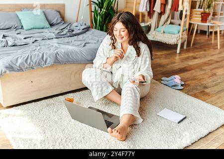 Eine junge Frau sitzt bequem auf einem weichen Teppich, nimmt ihren Laptop und schlürft Tee. Stockfoto