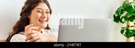 Eine glückliche Frau in Übergröße lächelt, während sie ihren Laptop in einem gemütlichen Raum benutzt. Stockfoto