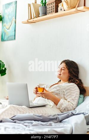 Eine junge, schöne Frau in Übergröße entspannt sich in ihrem gemütlichen Zuhause und schlürft Tee, während sie ihren Laptop benutzt. Stockfoto