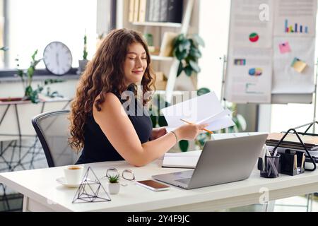 Eine junge Frau in Übergröße mit langen Locken konzentriert sich auf die Arbeit in ihrem Büro, indem sie ihre Aufgaben kreativ und selbstbewusst ausgleicht. Stockfoto