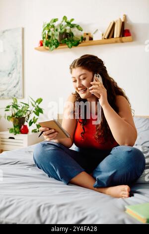 Eine fröhliche Frau in Übergröße sitzt bequem auf ihrem Bett, führt ein lebhaftes Telefongespräch und schreibt sich Notizen. Stockfoto