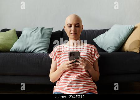 Eine junge Frau mit Alopezie sitzt bequem auf einer Couch, in ihr Smartphone eingesogen. Stockfoto