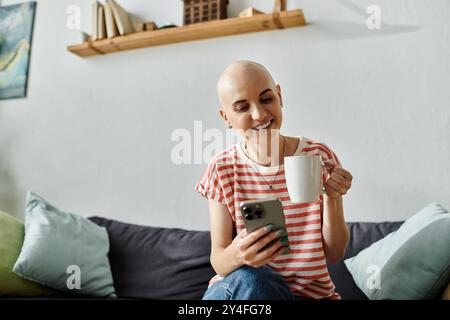 Eine junge Frau mit Alopezie lächelt, während sie aus einem Becher schlürft und ihr Telefon überprüft. Stockfoto