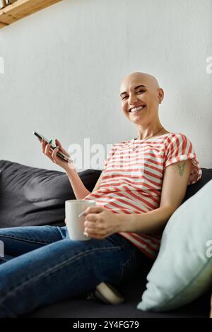 Eine lächelnde kahlköpfige Frau entspannt sich auf der Couch, hält eine Tasse und blättert auf ihrem Handy. Stockfoto