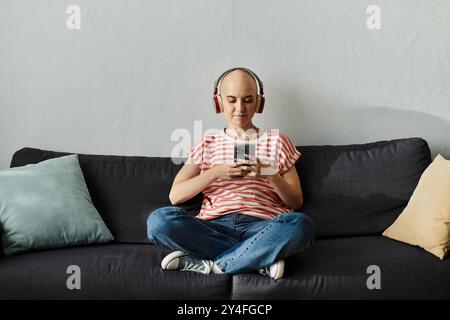 Eine junge, kahlköpfige Frau sitzt bequem auf einer Couch, verloren in ihrer Musik mit Kopfhörern. Stockfoto