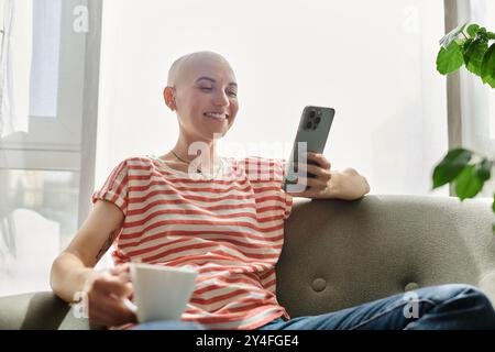 Eine kahlköpfige Frau mit Alopezie lächelt sanft, während sie sich mit einer Tasse Kaffee und ihrem Telefon entspannt. Stockfoto