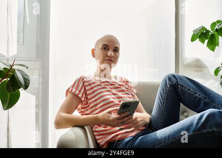 Eine junge kahlköpfige Frau mit Alopezie sitzt bequem auf einem Sofa und greift mit ihrem Smartphone auf. Stockfoto