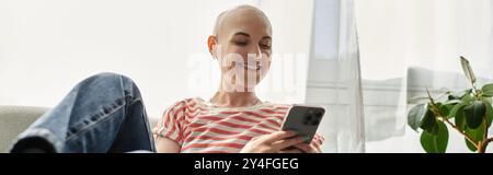 Eine junge kahlköpfige Frau mit Alopezie lächelt, während sie zu Hause ihr Smartphone bedient. Stockfoto