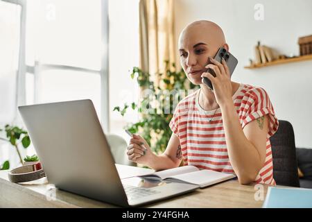 Eine junge Frau mit Alopezie lächelt, während sie am Telefon spricht, und taucht in ihr Heimbüro ein. Stockfoto