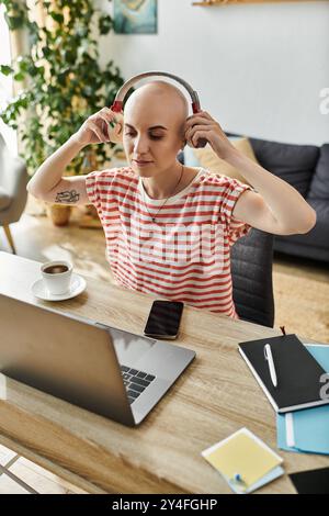 Eine schöne kahlköpfige Frau sitzt bequem, konzentriert auf ihre Arbeit, trägt Kopfhörer und schlürft Kaffee. Stockfoto