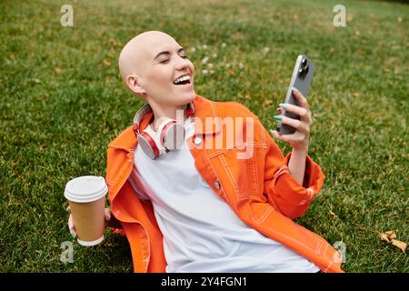 Eine fröhliche kahlköpfige Frau entspannt sich auf dem Gras, schlürft Kaffee und lächelt an ihrem Handy. Stockfoto