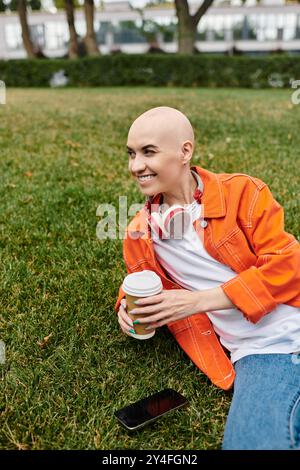 Eine junge Frau mit Alopezie lächelt, während sie eine Kaffeetasse auf dem Gras hält und Wärme ausstrahlt. Stockfoto