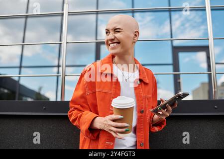 Eine junge kahlköpfige Frau genießt einen sonnigen Tag, schlürft Kaffee und schreibt auf ihrem Telefon, verstrahlt Freude. Stockfoto