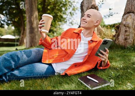 Eine fröhliche junge Frau entspannt sich auf dem Gras und trinkt Kaffee, während sie ihr Smartphone benutzt. Stockfoto