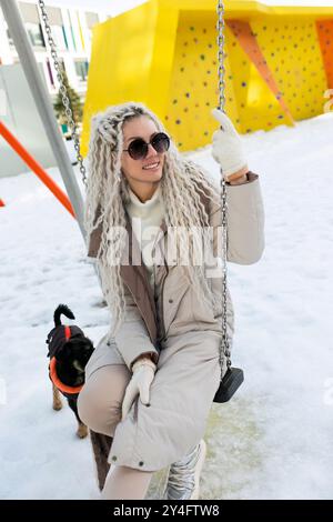 Eine Frau sitzt auf einer schneebedeckten Schaukel, gebündelt in warmer Kleidung. Der schneebedeckte Boden steht im Kontrast zu ihrem dunklen Outfit, während sie besinnlich aussieht Stockfoto