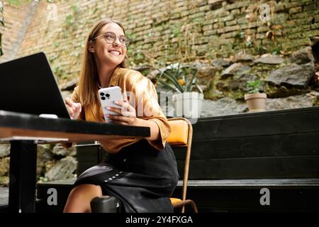 In einem lebhaften Café greift eine junge Frau mit einer Beinprothese glücklich mit ihrem Smartphone und Laptop. Stockfoto