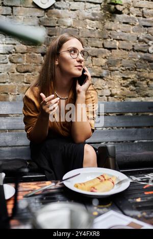 Eine junge Frau mit einem künstlichen Bein führt ein lebhaftes Telefongespräch, während sie in einem malerischen Café eine Mahlzeit genießt. Stockfoto