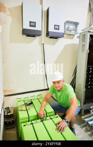 Porträt des Technikers, der am grünen Batteriespeicher auf dem Boden arbeitet. An der Wand zwei weiße moderne Wechselrichtereinheiten, die über schwarze Kabel mit der Batterie verbunden sind. Mann, der das Sonnenkollektorsystem installiert. Stockfoto