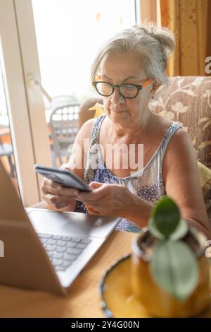 Eine ältere Frau sitzt zu Hause und konzentriert sich auf ihr Smartphone mit einem Laptop vor sich. Stockfoto