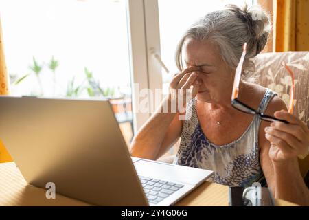 Eine ältere Frau hält ihre Brille und reibt sich die Augen, nachdem sie zu Hause an einem Laptop gearbeitet hat. Stockfoto