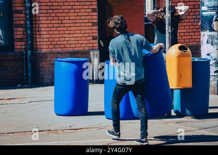 Ein junger Mann trägt ein großes blaues Fass, mehrere blaue Fässer im Hintergrund, Gehweg, von hinten fotografiert Stockfoto