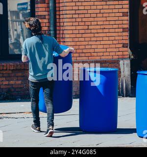 Ein junger Mann trägt ein großes blaues Fass, mehrere blaue Fässer im Hintergrund, Gehweg, von hinten fotografiert Stockfoto