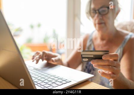 Eine ältere Frau tippt auf ihrem Laptop, während sie eine Kreditkarte hält und eine Online-Zahlung von zu Hause aus durchführt. Stockfoto