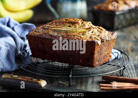 Vorderseite: Frisches, hausgemachtes Bananenbrot, das auf einem Bäckerregal mit Zutaten in der Nähe gekühlt wird. Selektiver Fokus auf Brotlaib mit verschwommenem Hintergrund Stockfoto