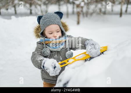 Ein entzückender kleiner Junge, der hilft, einen Schnee aus einem Auto zu bürsten. Mommy ist kleine Helferin. Winteraktivitäten für kleine Kinder. Stockfoto