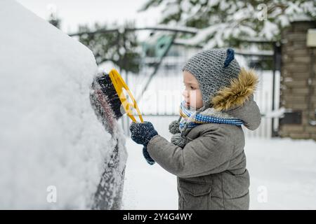 Ein entzückender kleiner Junge, der hilft, einen Schnee aus einem Auto zu bürsten. Mommy ist kleine Helferin. Winteraktivitäten für kleine Kinder. Stockfoto