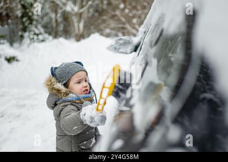 Ein entzückender kleiner Junge, der hilft, einen Schnee aus einem Auto zu bürsten. Mommy ist kleine Helferin. Winteraktivitäten für kleine Kinder. Stockfoto