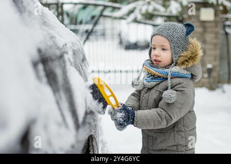 Ein entzückender kleiner Junge, der hilft, einen Schnee aus einem Auto zu bürsten. Mommy ist kleine Helferin. Winteraktivitäten für kleine Kinder. Stockfoto