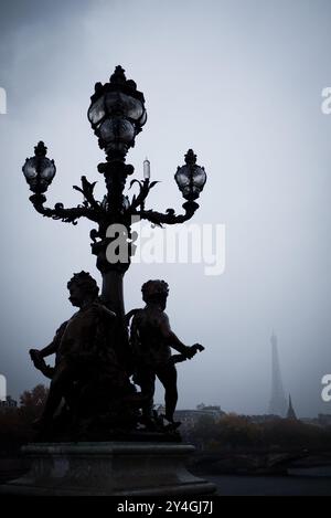 Laterne mit Engelchen auf der Pont Alexandre III an einem regnerischen, bewölkten Tag mit dem Eiffelturm in Paris, Frankreich | PARIS, Frankreich — Bronzene Engelchen krönen eine verzierte Laterne auf der Pont Alexandre III, die sich vor einem regnerischen Himmel abzeichnet, während der Eiffelturm im Nebel dahinter verschwindet. Die Brücke wurde für die Weltausstellung 1900 eröffnet und ist bekannt für ihr Art-Nouveau-Straßenmobiliar, das den Grand Palais und den Petit Palais mit Les Invalides verbindet. Das diffuse Licht drückt beide Strukturen in gleichmäßige graublaue Töne. Stockfoto
