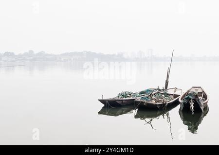 HANOI, Vietnam – drei mit Fischernetzen beladene hölzerne Sampane liegen am West Lake (Ho Tay) in Hanoi, umgeben von starkem Dunst. Die traditionellen Boote stehen im Kontrast zum modernen Stadtbild und bieten einen Einblick in Vietnams dauerhafte Fischerkultur inmitten städtischer Entwicklung und ökologischer Herausforderungen. Stockfoto