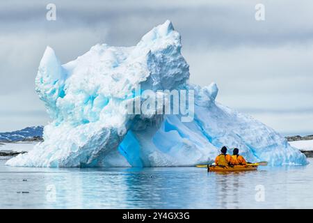 Ein Paar Kajakfahrer paddeln in einem Tandem-Kajak in der Nähe eines großen, dramatisch geschnitzten blauen Eisbergs auf Petermann Island auf der Antarktischen Halbinsel. Stockfoto