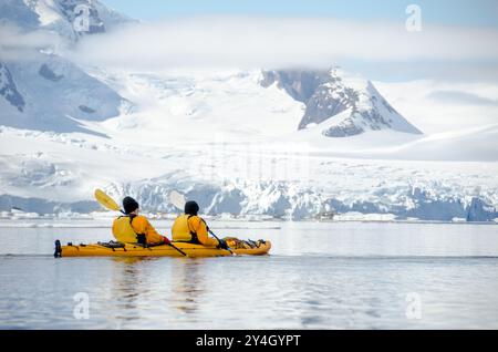 Petermann Island Sea Kayak Antarktische Halbinsel Gletscher / PETERMANN ISLAND, Antarktis — zwei Kajakfahrer paddeln mit einem gelben Tandem-Kajak über das spiegelruhige Wasser vor Petermann Island im Wilhelm-Archipel, hinter dem sich Gletscher-bedeckte Gipfel der westlichen Antarktischen Halbinsel erheben. Petermann Island liegt südlich des Lemaire-Kanals und beherbergt Gentoo- und Adélie-Pinguin-Kolonien, die sich in entgegengesetzte Richtungen bewegen – Gentoo-Zahlen stiegen ungefähr 27 % an, während Adélie-Zahlen zwischen 2003/04 und 2007/08 ungefähr 29 % fielen, als die Halbinsel erwärmt wurde. Stockfoto