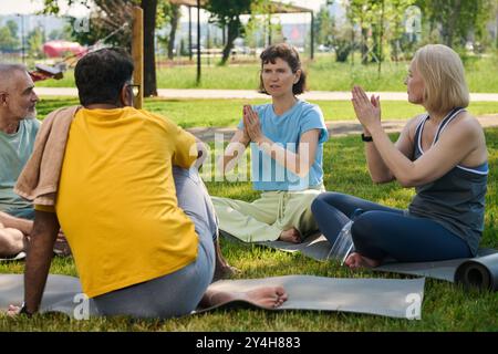Zwei reife Frauen halten die Hände zusammen, während sie vor interkulturellen älteren Männern auf Matten sitzen und neue Bewegung zeigen Stockfoto