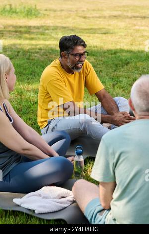 Ein glücklicher, multiethnischer reifer Mann in Activewear erzählt seine Geschichte mit einer Gruppe von Menschen, die auf Matten um ihn herum sitzen und die Pause genießen Stockfoto