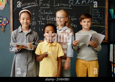 Porträt von vier jungen Schülern mit unterschiedlichem ethnischen Hintergrund, die fröhlich in die Kamera lächeln, während sie im Grundschulzimmer an der Tafel stehen Stockfoto