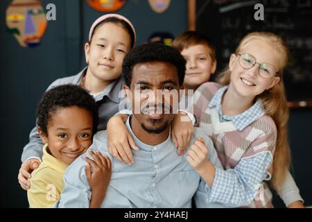 Porträt eines lächelnden afroamerikanischen Lehrers, der mit vier jüngeren Schülern posiert und einen glücklichen Lehrer im Klassenzimmer umarmt, um eine fröhliche Atmosphäre zu schaffen Stockfoto