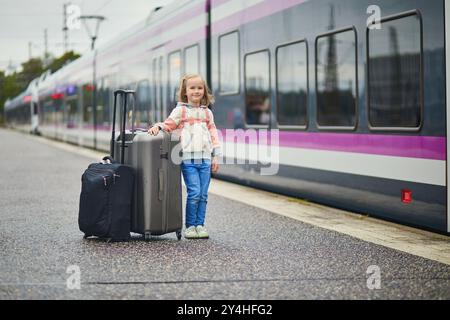 Entzückendes Mädchen im Vorschulalter auf einem Bahnhof. Kleines Kind, das auf einem Bahnsteig auf einen Zug wartet. Kind mit Gepäck bereit zu reisen. In den Urlaub Stockfoto