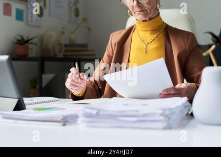Leitende Geschäftsfrau, die Dokumente hält, während sie in einer modernen Büroumgebung mit Brille am Schreibtisch sitzt und sich auf die Prüfung von Papieren konzentriert Stockfoto